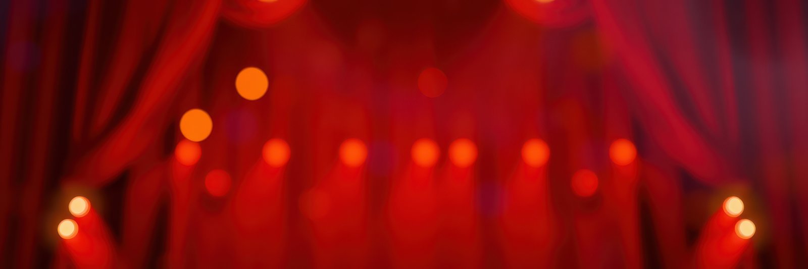 Bright stage lights illuminate a dramatic red curtain for an upcoming theatrical performance in the evening